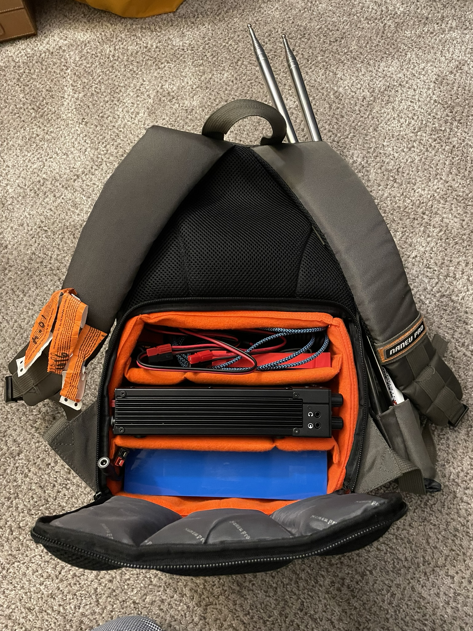 The backside of a backpack. A stealth panel is opened, showing an orange padded square with three vertically stacked compartments. The bottom compartment contains a battery, the middle compartment contains the Xiegu G90 transceiver. The upper compartment contains several wires. Two telescoping antenna whips are visible sticking up from the frontside.